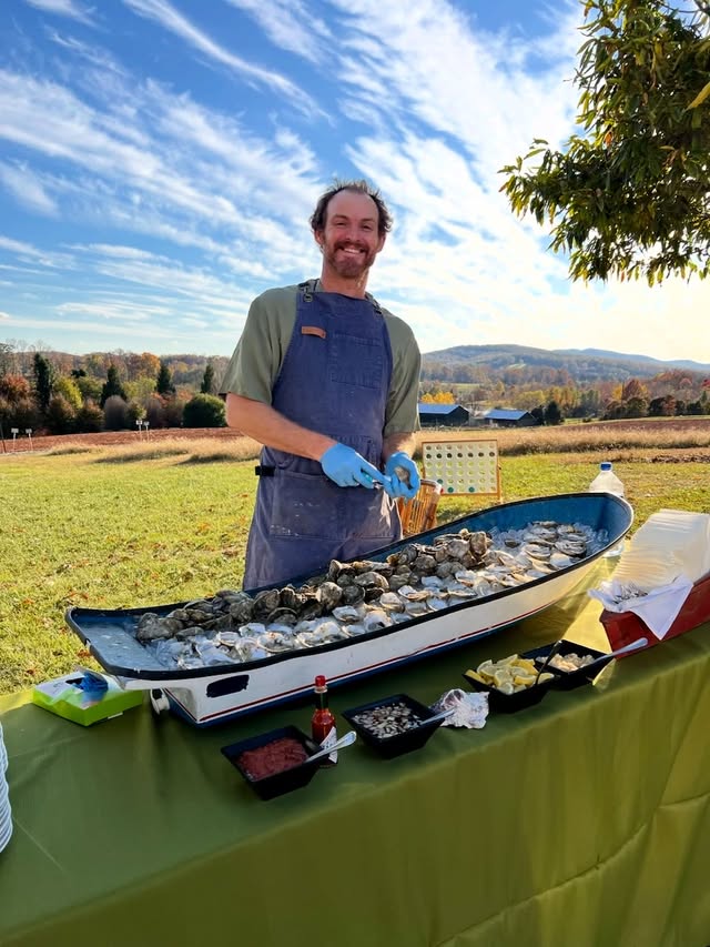 Eli shucking Revival oysters behind a boat-shaped raw bar full of ice, the Blue Ridge Mountains behind him